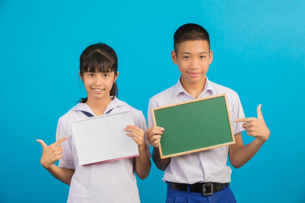 Two Asian students in school uniforms smiling while holding a notebook and a green board against a blue background