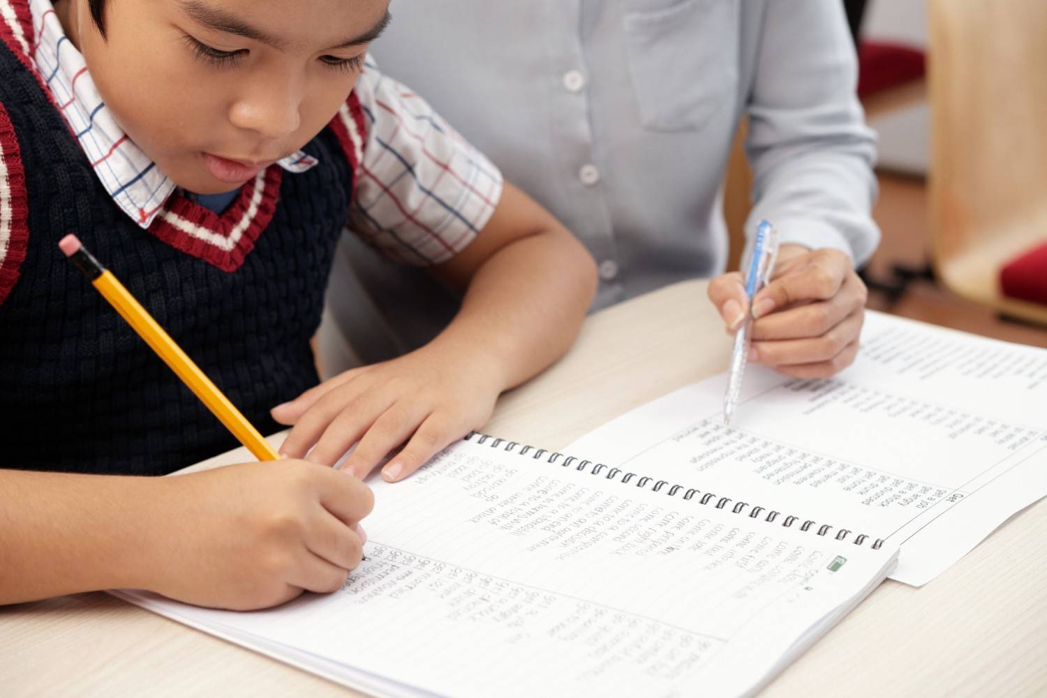 Asian schoolboy writing in notebook with teacher sitting beside him during tutoring session