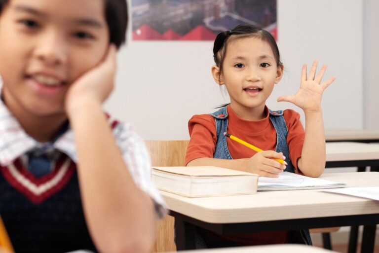 Young Asian girl raising hand to answer question while sitting at desk in classroom