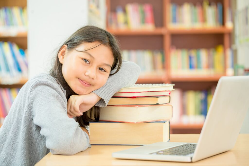 Student resting on a stack of books beside a laptop in the library