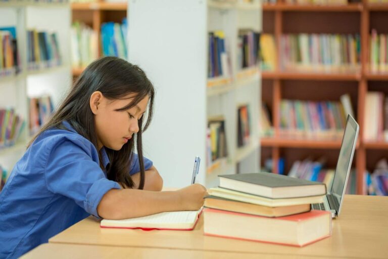 Student writing notes while studying in the library with books and laptop